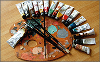 Four paintbrushes atop a used paint palette that is surrounded by paint tubes.