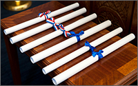 Six rolled up degrees, three tied with red-white-and-blue bows and three tied with plain blue bows, are in a row on a wooden display.