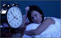 A woman laying stomach-down in bed, clutching her head, and staring at an alarm clock reading 3:50.