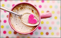 A top-down view of a spoon lifting a pink paper heart out of a mug filled with coffee.