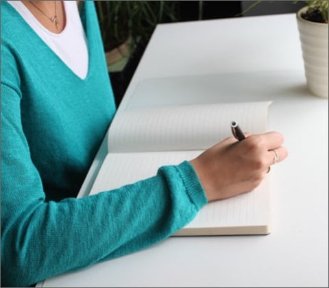 A woman wearing a teal sweater and writing in a notebook.